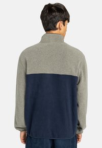 Fleece mikina s vysokým límcem, která má světlou šedou horní část a tmavě modrou dolní část. Měkká textura a volný střih.