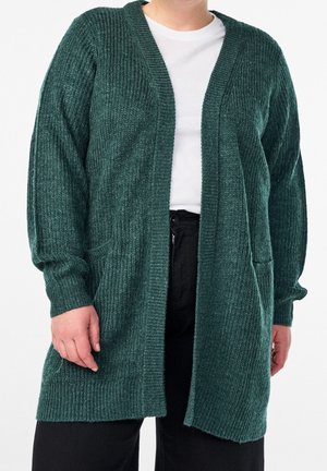Personne portant un cardigan en maille vert foncé ouvert devant, par-dessus une chemise blanche et un pantalon noir, sur un fond uni.