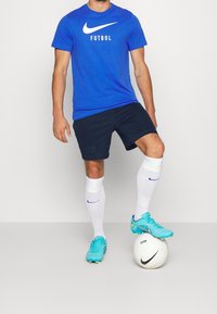 Camiseta azul de Nike con el texto "FUTBOL", pantalones cortos azul marino, calcetines blancos y botas de fútbol turquesa, de pie sobre un balón de fútbol blanco.
