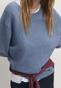 Maglione lavorato a maglia color azzurro chiaro con scollo rotondo e spalle scese, indossato sopra una maglietta bianca, abbinato a un accento cremisi annodato.