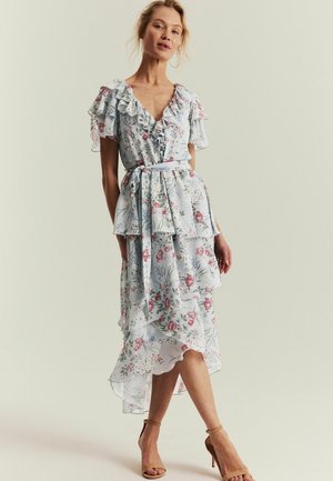 Frau trägt ein hellblaues, geblümtes Wickelkleid mit Rüschen, einem Taillengürtel und einem gestuften Rock und posiert vor einem schlichten Hintergrund.