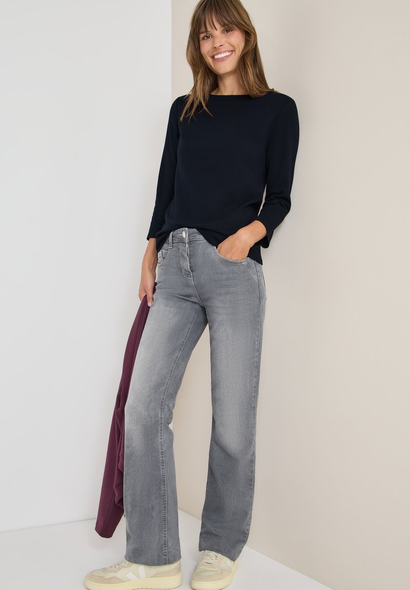 Jeans grigi larghi con vita media, abbinati a una maglietta a maniche lunghe blu scuro. Il modello è in piedi contro uno sfondo chiaro, mentre tiene una giacca bordeaux.