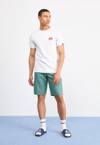 Weißes T-Shirt mit kurzen Ärmeln und buntem Logo, grüne Shorts, weiße Socken mit schwarzen Streifen und marineblaue Schlappen, stehend auf blauem Boden.