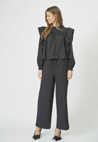 Blusa nera con spalline arricciate, vestibilità ampia e maniche lunghe. Abbinata a pantaloni neri larghi e tacchi a punta con dettagli a fiocco.