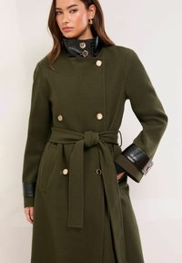 Płaszcz oliwkowozielony o kroju dwurzędowym, z czarnymi akcentami z imitacji skóry na mankietach i kołnierzu, przewiązany paskiem w talii oraz ze złotymi guzikami.