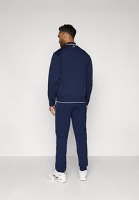 Σκούρο μπλε φούτερ και παντελόνια φόρμας από μαλακό ύφασμα, με ριγέ τελείωμα, λευκές λεπτομέρειες και ένα μικρό λογότυπο στον αυχένα.