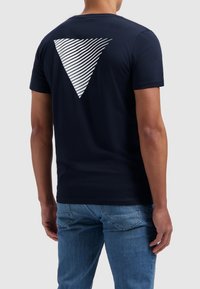 Marineblauwe katoenen T-shirt met een witte gestreepte driehoekige ontwerp op de rug. Korte mouwen en een klassieke pasvorm. G gedragen met lichtblauwe jeans.