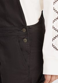 Mustad mustad denim üleõlad on varustatud küljetasku, trukk-nuppude ja siledaga tekstuuriga. Rõivast on kombineeritud valge pika varrukaga särgiga.