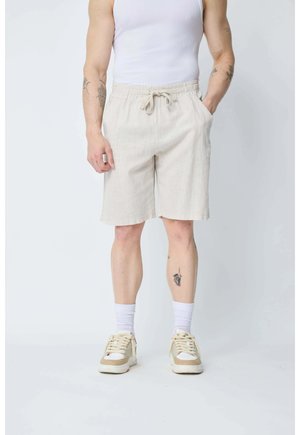 Homme portant un short beige à cordon, un débardeur blanc, des chaussettes blanches et des baskets beige, debout avec une main dans la poche.