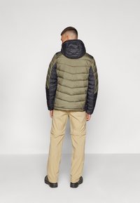 Padded jacket w oliwkowym korpusie, czarnych rękawach i kapturze. Posiada pikowany design i trwały materiał. Noszony z beżowymi spodniami i czarnymi butami.