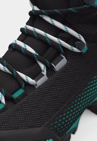 Schwarz-türkiser Sneaker mit strukturiertem Stoff, schwarz-weiß gemusterten Schnürsenkeln und Schlaufenösen, Nahaufnahme von Vorder- und Seitenansicht.