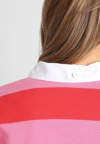 Sweter w paski z różowymi i czerwonymi pasami, z białym kołnierzykiem i zapięciem na guziki z tyłu. Widoczna tekstura dzianiny.