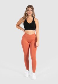 Schwarzer Sport-BH und leggings in Rostfarbe aus dehnbarem, glattem Stoff. Das Model trägt weiße Turnschuhe und steht mit neutralem Gesichtsausdruck.