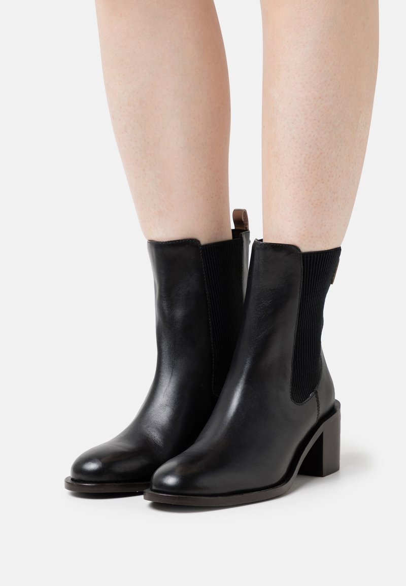 Botas de tobillo negras de cuero con paneles laterales elásticos, puntera cuadrada y tacón ancho. Textura suave con un brillo sutil y un diseño minimalista.