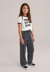 White t-shirt with "WHO LOT OF LUCK!" in black, paired with black floral-patterned pants. Canvas sneakers with black and white accents.