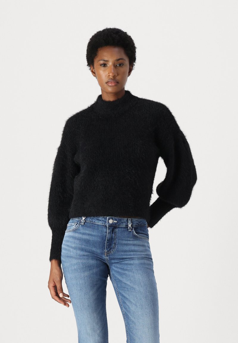 Maglione nero in maglia pelosa con collo alto, spalle cadenti e maniche a palloncino. Abbinato a jeans in denim blu.