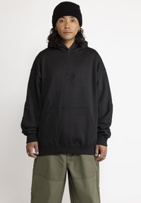 Zwarte hoodie met een voorzak, voorzien van geborduurde ontwerpen. Gemaakt van zachte stof, heeft het een relaxte pasvorm en ribgebreide manchetten. Gepaard met olijfgroene pantalons.