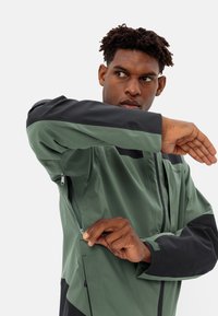 Green and black jacket with a zippered pocket, matte finish, adjustable cuffs, and structured panels for a streamlined look.