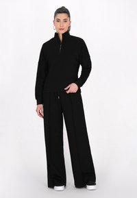 Felpa nera con zip e colletto alto, abbinata a pantaloni neri larghi. Entrambi presentano texture morbide e una silhouette rilassata.