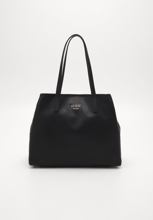 Zwarte leren Guess tote tas met twee lange handvatten en klein metalen logo aan de voorkant tegen witte achtergrond.