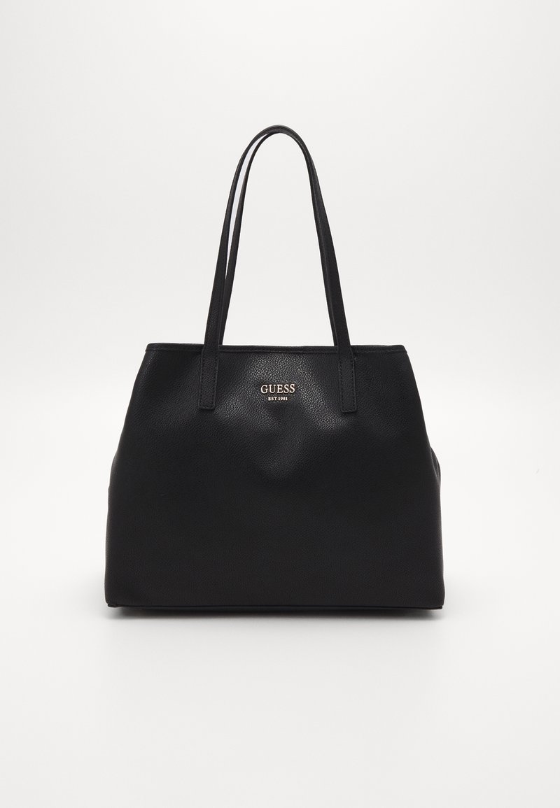 Sac tote Guess en cuir noir avec deux longues poignées et petit logo métallique sur le devant sur fond blanc.