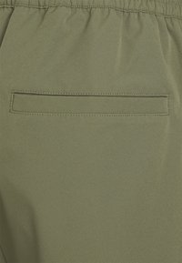 Tissu vert olive avec une texture lisse, comportant une poche arrière avec une ouverture plate. Coutures visibles autour de la zone de la poche.