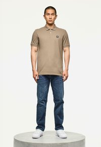 Man staat op een rond platform en draagt een bruine polo, blauwe jeans en witte sneakers tegen een effen lichte achtergrond.