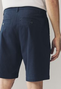 Tumesinised puuvillased lühikesed püksid siledast kangast, kahel tagataskul on nööpkinnitused, ning need on kitsas lõikega. Äär ulatub veidi üle põlve.