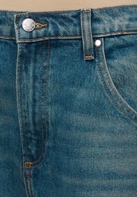 Κοντινό πλάνο μπλε τζιν με ζώνη μέσης, κουμπί, θηλιά ζώνης και μπροστινή τσέπη με ορατές πορτοκαλί ραφές.