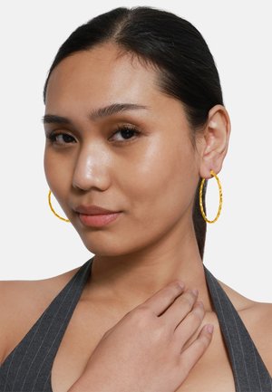 Goldfarbene, gedrehte Creolen. Runde Form mit strukturierter Oberfläche. Modell trägt ein graues Halsband-Top, Hand auf der Schulter.