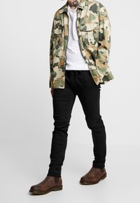Jas met camouflagepatroon in beige en groen, witte t-shirt, zwart taps toelopende broek, bruine enkellaarsjes, staande op een neutrale achtergrond.