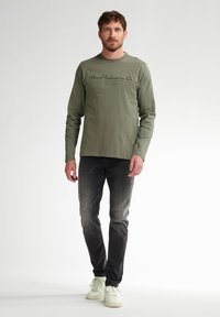 Cămașă verde măsliniu cu mâneci lungi și un logo tipărit negru, asociată cu blugi gri închis și adidași albi, într-o poziție standard, pe un fundal neutru.