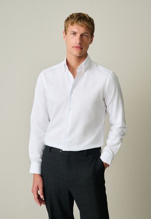 Hombre rubio con una camisa blanca de manga larga abotonada y pantalones oscuros a cuadros, de pie con una mano en el bolsillo.