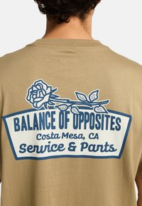 Tan T-shirt met een gedrukte grafiek van een roos en tekst: "BALANCE OF OPPOSITES, Costa Mesa, CA, Service & Parts" in marineblauw.