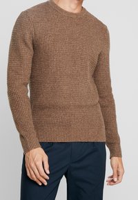 Man i en brun strukturerad stickad tröja och mörkblå byxor, stående med avslappnade armar vid sidorna mot en enfärgad bakgrund.