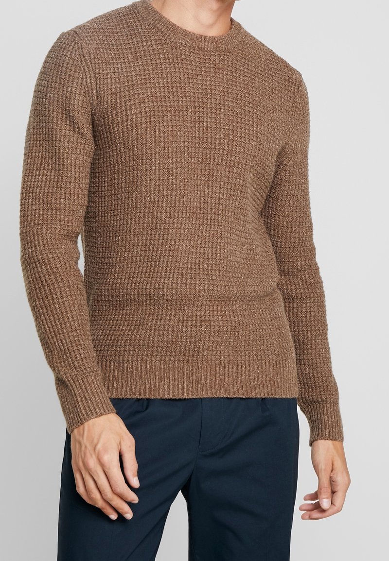 Man i en brun strukturerad stickad tröja och mörkblå byxor, stående med avslappnade armar vid sidorna mot en enfärgad bakgrund.