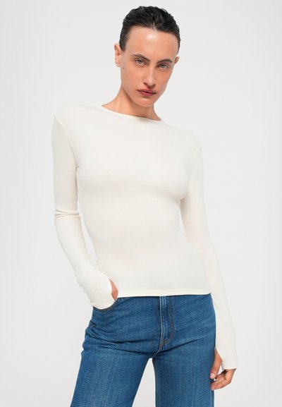 White long-sleeve top, fitted, made from a smooth fabric. Crew neckline, with extended sleeve cuffs. Paired with dark blue denim jeans.