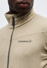 Άνδρας που φοράει μπεζ φλις ζακέτα με φερμουάρ και ψηλό γιακά, με το λογότυπο "NORRØNA" κεντημένο στο στήθος.