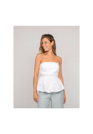 Femme souriante regardant sur le côté, portant un haut péplum blanc sans bretelles et un pantalon gris clair sur un fond uni.