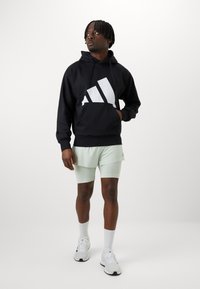 Juodas gobtuvas su dideliu baltu Adidas logotipu, derinamas su mėtų žalio spalvos šortais. Modelis dėvi baltus sportinius batelius ir iki kulkšnies siekiančias kojines.