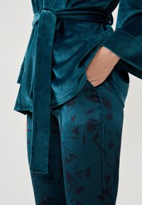 Top envolvente de terciopelo en color verde azulado con un cinturón de lazo; pantalones a juego en verde azulado con un patrón floral oscuro, que resaltan texturas suaves y líneas fluidas.