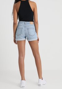Pantalones cortos de mezclilla azul claro con dobladillo enrollado, dos bolsillos traseros y una etiqueta de marca marrón en la cinturilla. Combinados con una blusa sin mangas negra.