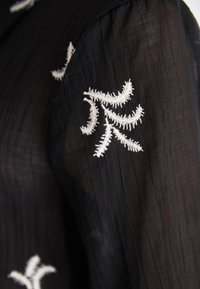 Bluză transparentă neagră cu broderie florală albă pe mânecă. Materialul are o textură ușor șifonată, care accentuează detaliile designului.