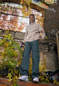 Veste zippée rose clair avec une finition texturée, associée à un pantalon cargo ample de couleur vert foncé avec de grandes poches latérales. Baskets blanches.