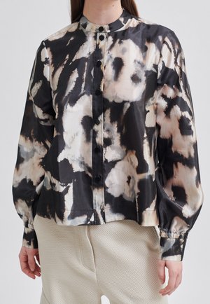 Žena oblečená v černé a béžové abstraktní blůze s dlouhými rukávy na knoflíky a texturované světle béžové kalhoty.