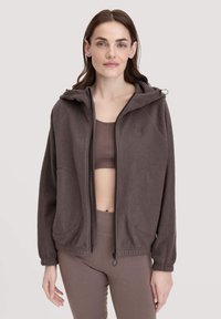 Dunkelbrauner Fleece-Reißverschluss-Hoodie mit Kapuze, zwei großen Taschen und gerippten Bündchen. Wird über einem passenden dunkelbraunen Oberteil getragen.