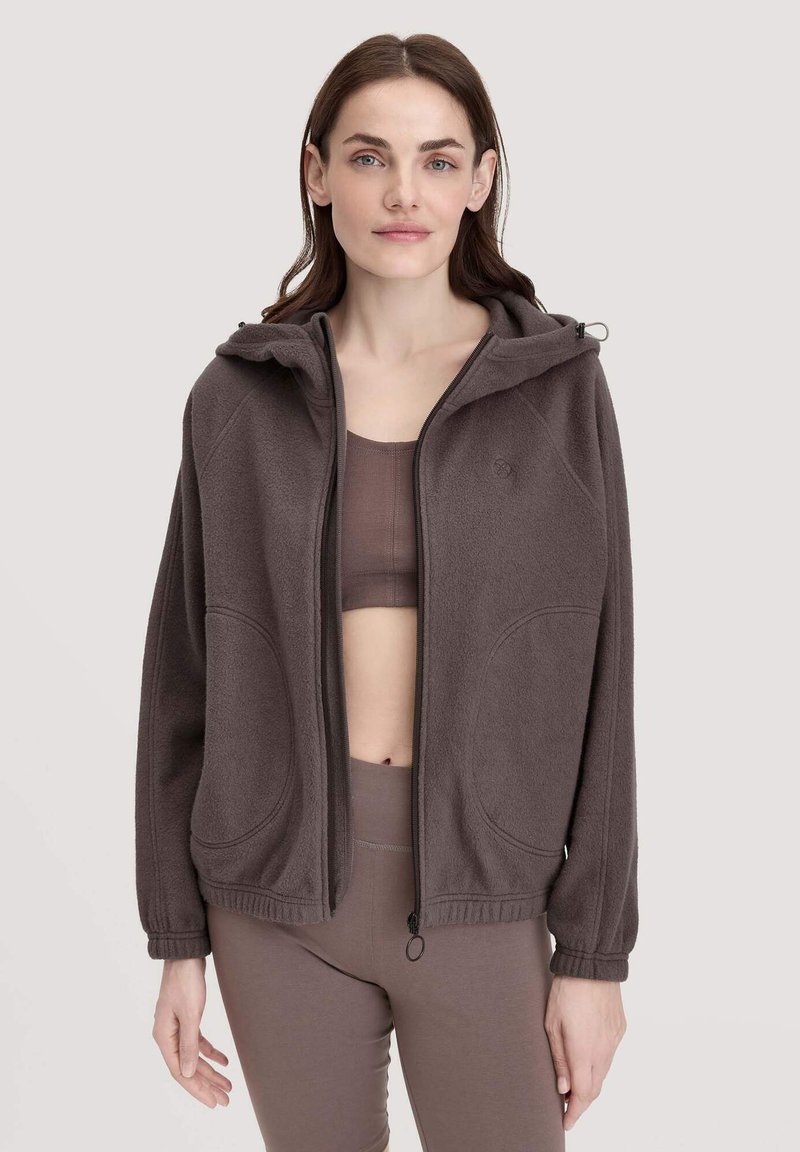 Dunkelbrauner Fleece-Reißverschluss-Hoodie mit Kapuze, zwei großen Taschen und gerippten Bündchen. Wird über einem passenden dunkelbraunen Oberteil getragen.