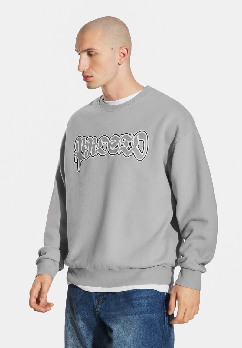 Jasnoszara bluza z okrągłym dekoltem i luźnym krojem. Na przodzie znajduje się wyraźna grafika w czerni i bieli. Miękka tekstura.