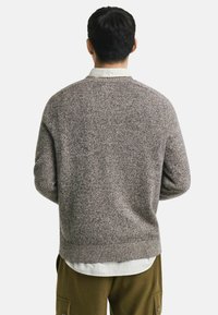 Braun gesprenkelter Strickpullover mit geripptem Saum, Rundhals und überschnittenen Schultern, getragen über einem weiß karierten Hemd mit grünen Akzenten.
