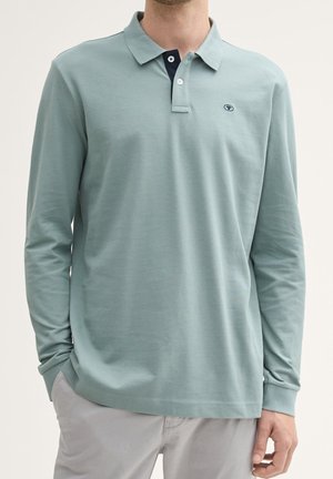 Homme portant un polo à manches longues vert clair avec un col à deux boutons et un petit logo brodé sur la poitrine, associé à un pantalon gris clair.